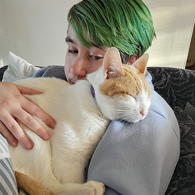 keeper is asleep on his owner's shoulder, who is gently holding him and gazing at him lovingly. His owner is a petite white person with short, green hair.