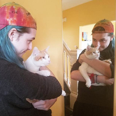 keeper is being held by his other owner, who has him in her arms and is standing in front of a mirror. keeper does not seem interested in his reflection. His owner is a tall white woman with long blue-green hair and a backwards baseball cap. She is smiling!