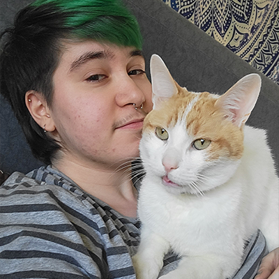 keeper lies on top of his owner's chest, with a small blep. His owner is a petite white person with short green hair, who is looking at the camera and smiling.